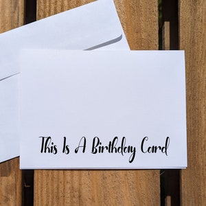 May include: A white card with black text that reads "This is a Birthday Card". The card is sitting on a wooden surface.