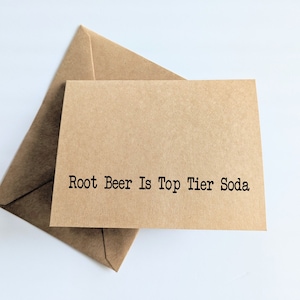 May include: A kraft paper greeting card with the text "Root Beer Is Top Tier Soda" in black typewriter font, resting on a matching envelope. The card and envelope are on a white background.