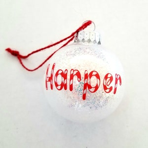 May include: White Christmas ornament with red glitter and the name "Harper" written in red candy cane font.