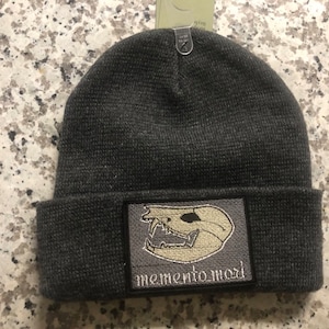 Puede incluir: Gorro gris con un parche negro que muestra una calavera blanca y el texto "memento mori".