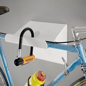 Op de afbeelding: Een blauwe fiets met een zwart U-slot en een witte wandfietsenrek. Het slot heeft een oranje en zwarte behuizing met de merknaam "Kryptonite". Een gele waterfles met een rode dop is ook zichtbaar.
