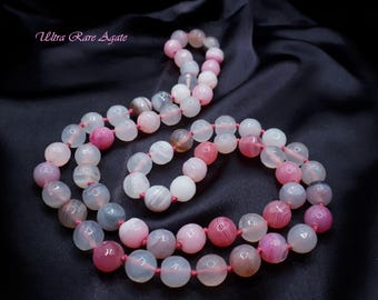 May include: A necklace of agate beads in shades of pink, white, and gray, strung on a red cord. The beads vary in size and pattern, with some displaying stripes and swirls. The text "Ultra Rare Agate" is in the upper left corner.