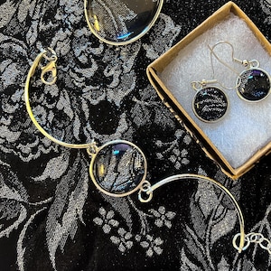 May include: A jewelry set featuring a silver bracelet, two oval pendants, and a pair of earrings. Each piece has a black background with a silver floral design. The earrings are in a small box.
