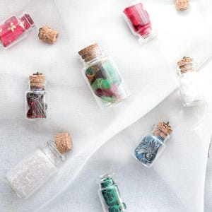 Puede incluir: Surtido de pequeñas botellas de vidrio con tapas de corcho, llenas de recortes de tela de colores. Las botellas están dispuestas sobre un fondo de tela blanca.