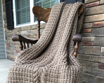 May include: A brown knitted throw blanket with a cable knit pattern draped over a wooden rocking chair.