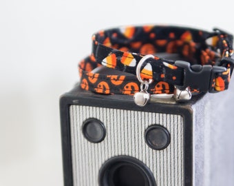 May include: A black and orange cat collar with a candy corn and ghost pattern. The collar has a silver bell and a black plastic buckle.