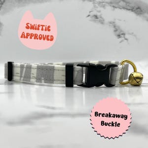 May include: A white and gray striped cat collar with a black breakaway buckle and a gold bell. The collar has the text "Swiftie Approved" on a pink cat-shaped tag.