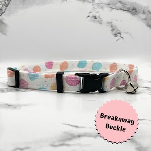 May include: White cat collar with a colorful cupcake pattern, featuring pink, orange, and blue accents. The collar has a black breakaway buckle and a silver bell. The text "Breakaway Buckle" is on a pink sticker.