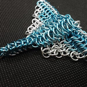 Könnte beinhalten: Ein handgefertigtes Kettenhemd-Stück in dreieckiger Form, bestehend aus ineinandergreifenden Metallringen. Das Design zeigt eine Kombination aus silbernen und leuchtend blauen Ringen, die einen auffälligen Kontrast erzeugen. Das Kettenhemd wird vor einem dunklen, strukturierten Hintergrund präsentiert.