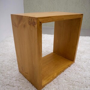 May include: A rectangular wooden side table with a natural light brown finish and visible wood grain. The piece features a hollow, open-frame construction with four flat sides, creating a simple, functional storage space within the center.