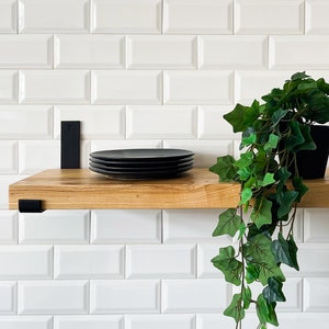May include: A wooden shelf with black metal brackets is mounted on a white metro tile wall. A stack of black plates sits on the shelf. A potted green plant hangs from the shelf.