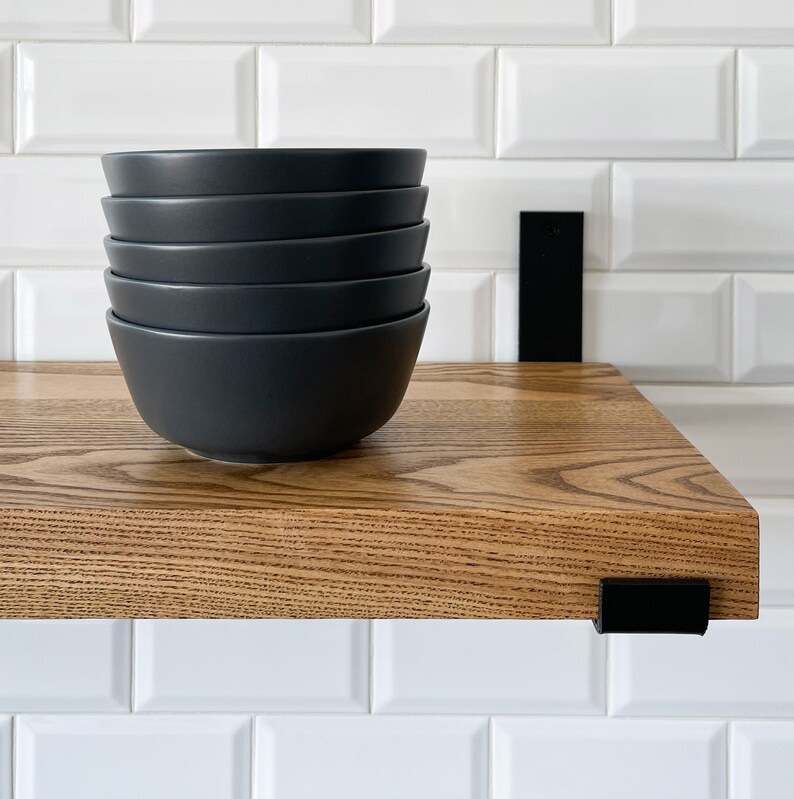 May include: A stack of five dark grey ceramic bowls on a wooden shelf with black metal brackets. The shelf is mounted to a white tiled wall.