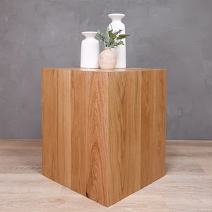 May include: A wooden cube-shaped end table with a light brown wood grain finish. The table is topped with three white ceramic vases, one of which holds a small bouquet of green leaves.