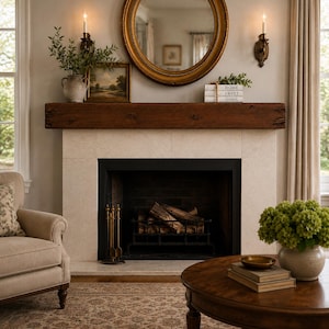 May include: A fireplace with a dark wood mantel, a cream-colored stone surround, and a black metal firebox containing logs. Above the mantel sits a round gold-framed mirror, two wall sconces, a framed landscape painting, and stacked books.