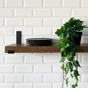 May include: A dark wood shelf with black metal brackets, mounted on a white metro tile wall. A stack of black plates sits on the shelf, and a potted green plant hangs over the edge.
