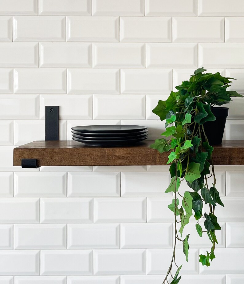 May include: A dark wood shelf with black metal brackets, mounted on a white metro tile wall. A stack of black plates sits on the shelf, and a potted green plant hangs over the edge.
