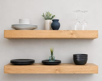 May include: Two horizontal light brown wooden floating shelves mounted on a wall. The top shelf holds a stack of white plates, a small potted green plant, a dark blue vase, and two clear glass wine glasses. The bottom shelf holds a stack of black plates, a small clear glass vase with a green plant, and a stack of black bowls.