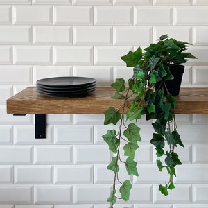 May include: A wooden shelf with black metal brackets, mounted on a white metro tile wall. A stack of black plates sits on the shelf, and a potted green plant hangs over the edge.