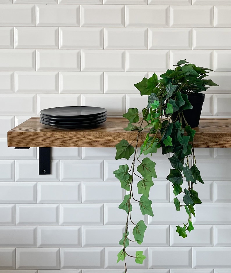 May include: A wooden shelf with black metal brackets, mounted on a white metro tile wall. A stack of black plates sits on the shelf, and a potted green plant hangs over the edge.