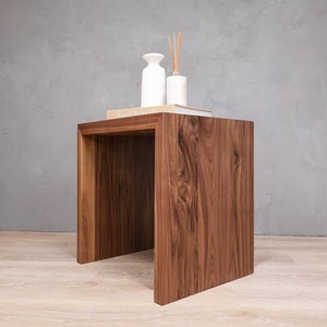 May include: A dark brown wooden side table with a square top and open sides. The table has a book titled "Houses Atelier AM" on top, along with a white vase and a diffuser.