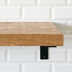 May include: A wooden shelf with a natural wood finish and black metal brackets. The shelf is mounted on a white tiled wall.