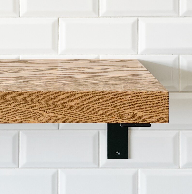 May include: A wooden shelf with a natural wood finish and black metal brackets. The shelf is mounted on a white tiled wall.