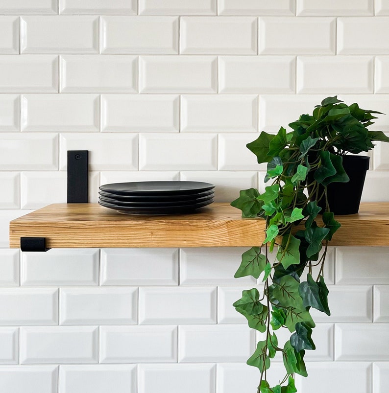 May include: A wooden shelf with black metal brackets is mounted on a white metro tile wall. A stack of black plates sits on the shelf. A potted green plant hangs from the shelf.