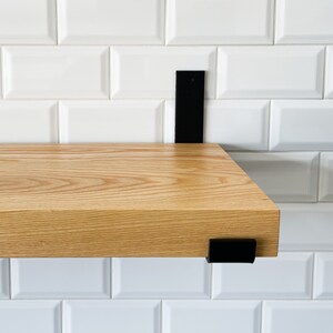 May include: A wooden shelf with black metal brackets mounted on a white tiled wall. The shelf is made of light wood with a natural grain pattern.
