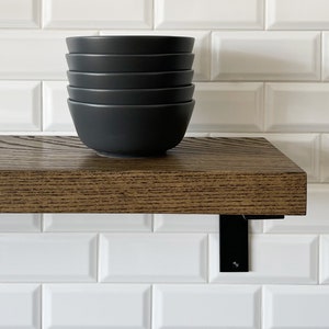 May include: A set of five black ceramic bowls stacked on a dark brown wooden shelf with black metal brackets. The shelf is mounted on a white metro tile wall.
