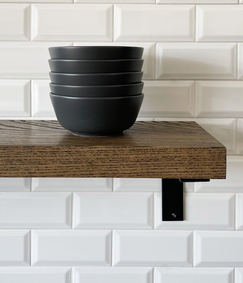 May include: A set of five black ceramic bowls stacked on a dark brown wooden shelf with black metal brackets. The shelf is mounted on a white metro tile wall.