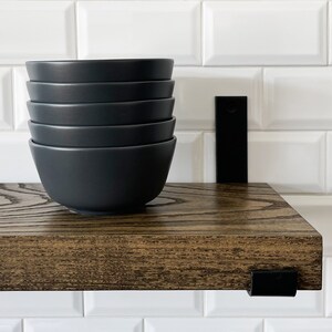 May include: A dark brown wooden shelf with black metal brackets. The shelf is mounted on a white tiled wall. Five black bowls are stacked on the shelf.