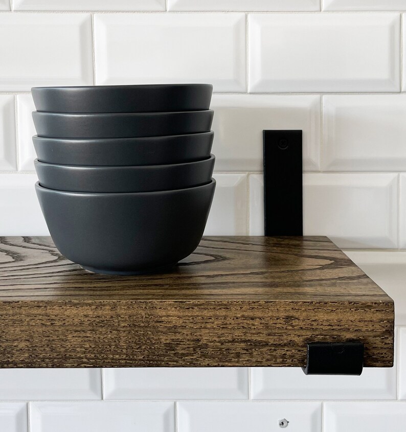May include: A dark brown wooden shelf with black metal brackets. The shelf is mounted on a white tiled wall. Five black bowls are stacked on the shelf.