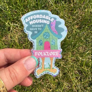 May include: A whimsical sticker with the text "AFFORDABLE HOUSING DOESN'T HAVE TO BE FOLKLORE." The sticker features a cartoon house with a green body, pink door, and a blue roof. The sticker has a glittery border and is held against a background of green grass.