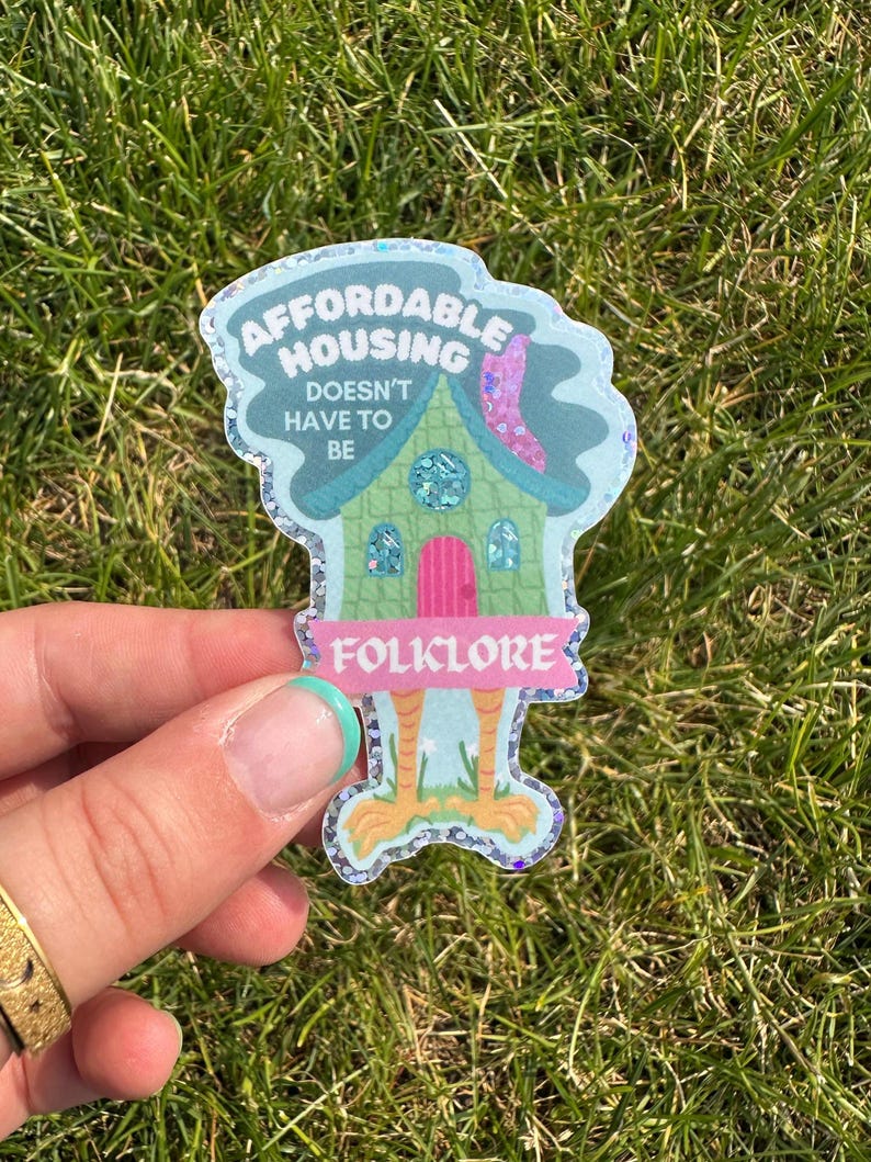 May include: A whimsical sticker with the text "AFFORDABLE HOUSING DOESN'T HAVE TO BE FOLKLORE." The sticker features a cartoon house with a green body, pink door, and a blue roof. The sticker has a glittery border and is held against a background of green grass.