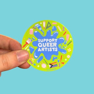 May include: A round, lime green sticker with the text "SUPPORT QUEER ARTISTS" in white and blue. The sticker features various art supplies and a rainbow flag design. The sticker is held by a hand.