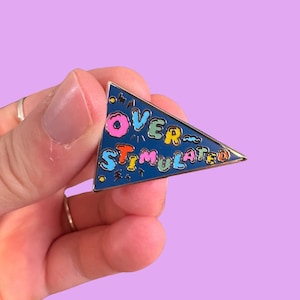 May include: A blue enamel pin in a pennant shape with the words "OVER STIMULATED" in colorful bubble letters. The pin has a gold border and is held against a purple background.