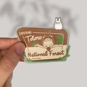 Puede incluir: Una pegatina verde y marr&oacute;n con un Totoro de dibujos animados sentado en una rama. La pegatina dice "Entering Totoro National Forest" con las palabras "Outlet Orion" en la parte inferior. Un peque&ntilde;o personaje de dibujos animados blanco est&aacute; en la parte superior del cartel.