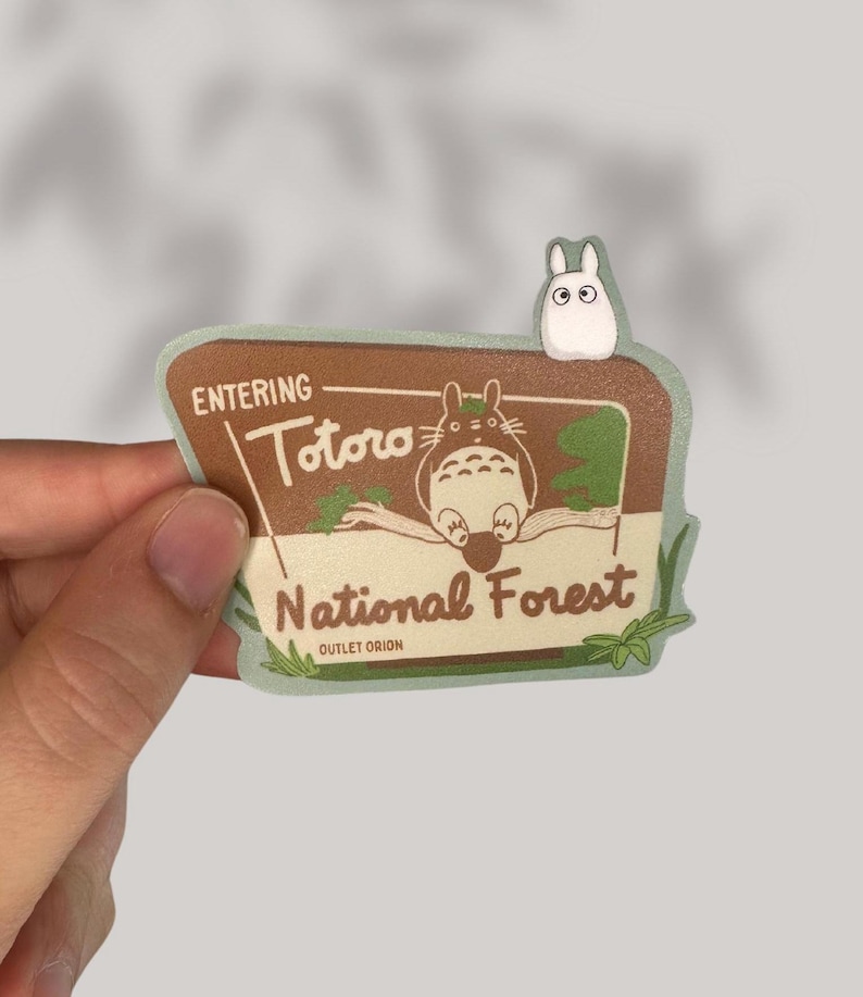 Puede incluir: Una pegatina verde y marr&oacute;n con un Totoro de dibujos animados sentado en una rama. La pegatina dice "Entering Totoro National Forest" con las palabras "Outlet Orion" en la parte inferior. Un peque&ntilde;o personaje de dibujos animados blanco est&aacute; en la parte superior del cartel.