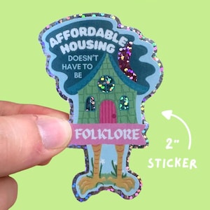 May include: A holographic glitter finish sticker with the text "AFFORDABLE HOUSING DOESN'T HAVE TO BE FOLKLORE". The sticker is shaped like a house with chicken legs and is approximately 2 inches in size.