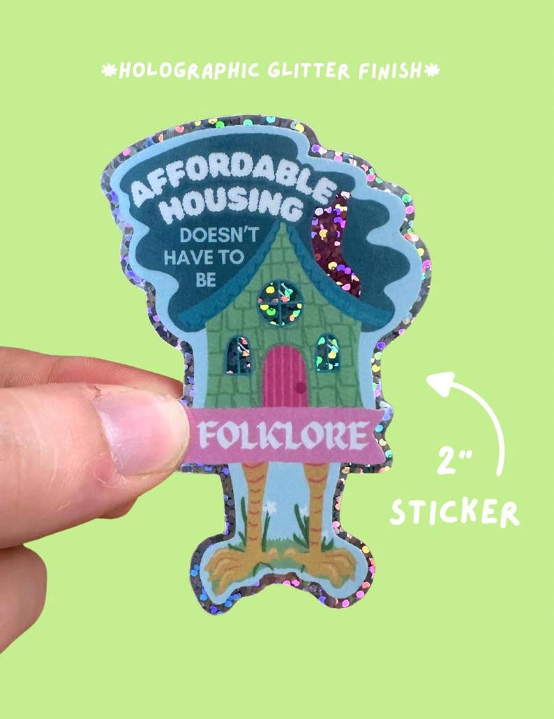 May include: A holographic glitter finish sticker with the text "AFFORDABLE HOUSING DOESN'T HAVE TO BE FOLKLORE". The sticker is shaped like a house with chicken legs and is approximately 2 inches in size.