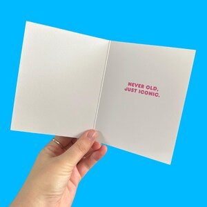 Puede incluir: Tarjeta de felicitaci&oacute;n blanca abierta con el texto rosa "NEVER OLD, JUST ICONIC". La tarjeta se sostiene sobre un fondo azul brillante, mostrando el mensaje interior.