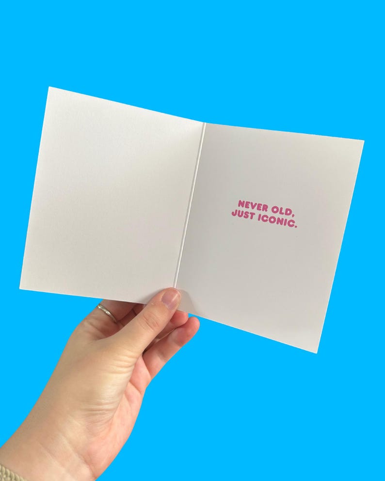 Puede incluir: Tarjeta de felicitaci&oacute;n blanca abierta con el texto rosa "NEVER OLD, JUST ICONIC". La tarjeta se sostiene sobre un fondo azul brillante, mostrando el mensaje interior.