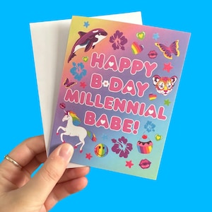 Puede incluir: Una colorida tarjeta de cumplea&ntilde;os con el texto "HAPPY B&bull;DAY MILLENNIAL BABE!" presenta un unicornio arco&iacute;ris, una orca y otros gr&aacute;ficos. La tarjeta se sostiene contra un sobre blanco, con un fondo azul brillante.