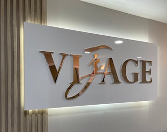 Puede incluir: Un letrero blanco con letras doradas que dice "VISAGE" con una figura femenina estilizada en la "A".