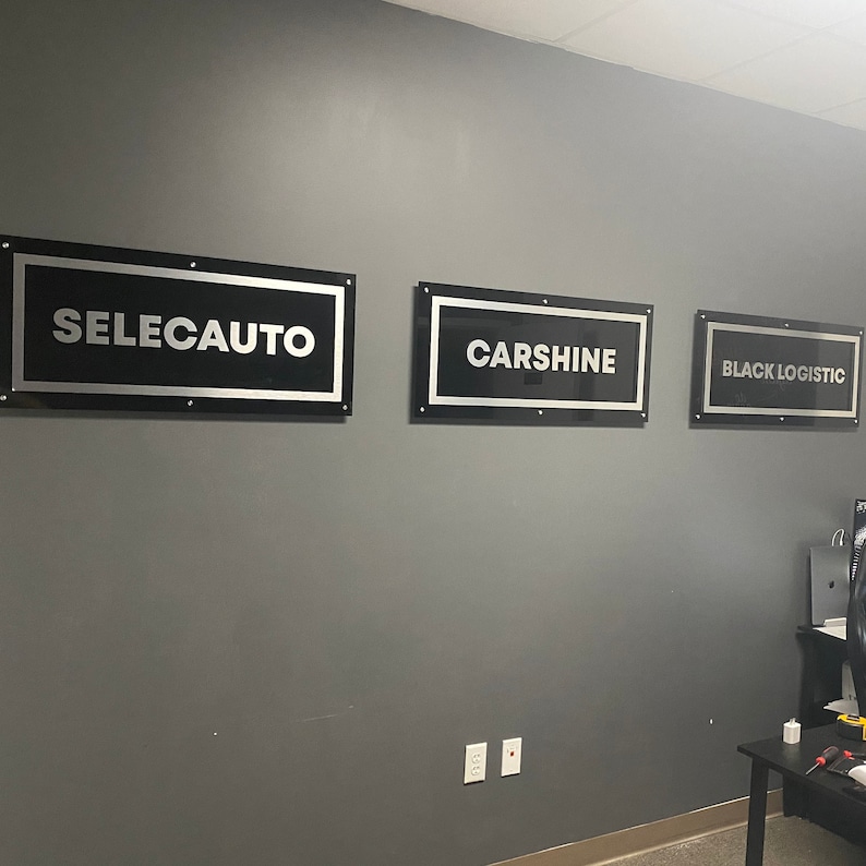 May include: Three black and silver rectangular signs with white text on a gray wall. The signs read "SELECAUTO", "CARSHINE", and "BLACK LOGISTIC".