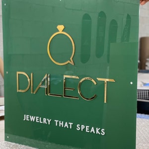 May include: Green sign with gold lettering that reads "Dialect Jewelry That Speaks". The logo is a gold ring with a diamond in the center.