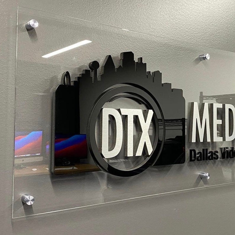 May include: A clear acrylic sign with a black and silver logo. The logo features a stylized camera with a cityscape inside the lens. The text "DTX MEDIA" and "Dallas Video" are visible on the sign.