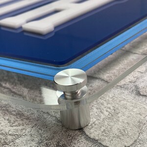 May include: Close-up of a silver metal stand with a round top and a cylindrical base. The stand is attached to a clear acrylic surface.