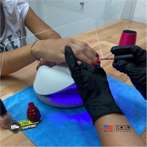 May include: A person is getting their nails done with a UV lamp and red nail polish. The person is wearing black gloves. The lamp is white with a blue light. There is a bottle of red nail polish and a bottle of clear nail polish on a blue towel.