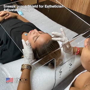 Könnte beinhalten: Klarer Sichtschutz aus Kunststoff für eine Kosmetikerin. Der Schutzschild ist höhenverstellbar und hat einen weißen Sockel. Der Text "Sneeze guard Shield for Esthetician Adjustable Height" ist auf dem Schild aufgedruckt. Der Text "BOLD DESIGNS Made in USA" ist auf dem Sockel aufgedruckt.